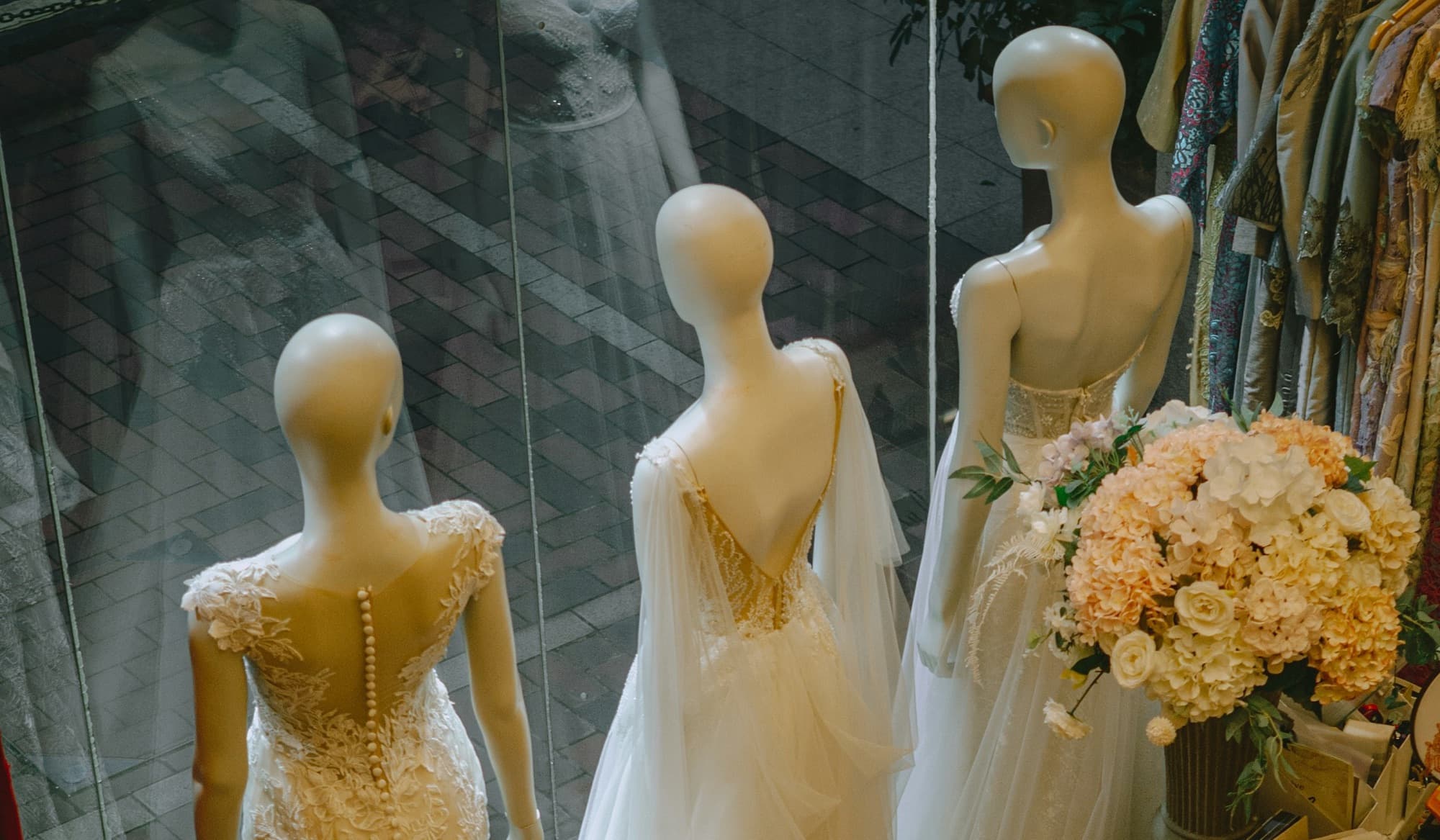Three mannequins dressed in ivory gowns in a boutique window, seen from above